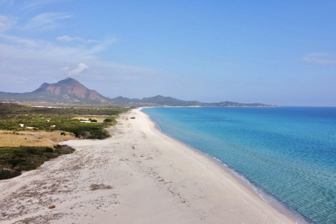 Paisaje costero con una playa de arena, aguas azules cristalinas y montañas a lo lejos.