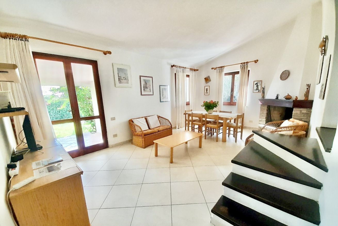Ein geräumiges Wohnzimmer mit einem Holztisch, einer Couch und einem Essbereich mit Stühlen. Ein Kamin und eine Treppe in eine weitere Ebene sind ebenfalls vorhanden.