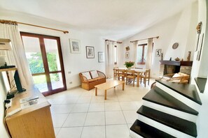 A spacious living room with a wooden coffee table, a sofa, and a dining area with chairs. There is a fireplace and a staircase leading to another level. - <i>Reclaim your own space on Sardinia's south-eastern coast, in a secluded, sheltered home a stone's throw from the sea and the heart of Costa Rei.</i>Just steps from the <b>centre of Costa Rei</b> and only <b>300 metres from the white sandy beach</b>, < (Muravera)
