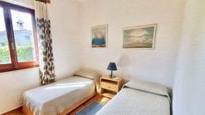 Ein Schlafzimmer mit zwei Einzelbetten, einem Nachttisch mit Lampe, einem Fenster mit Vorhängen und zwei Wandgemälden.