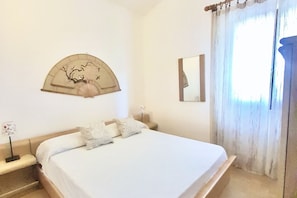 A bedroom with a bed, a wall-mounted fan, a mirror, and a window with curtains. - <i>Reclaim your own space on Sardinia's south-eastern coast, in a secluded, sheltered home a stone's throw from the sea and the heart of Costa Rei.</i>Just steps from the <b>centre of Costa Rei</b> and only <b>300 metres from the white sandy beach</b>, < (Muravera)