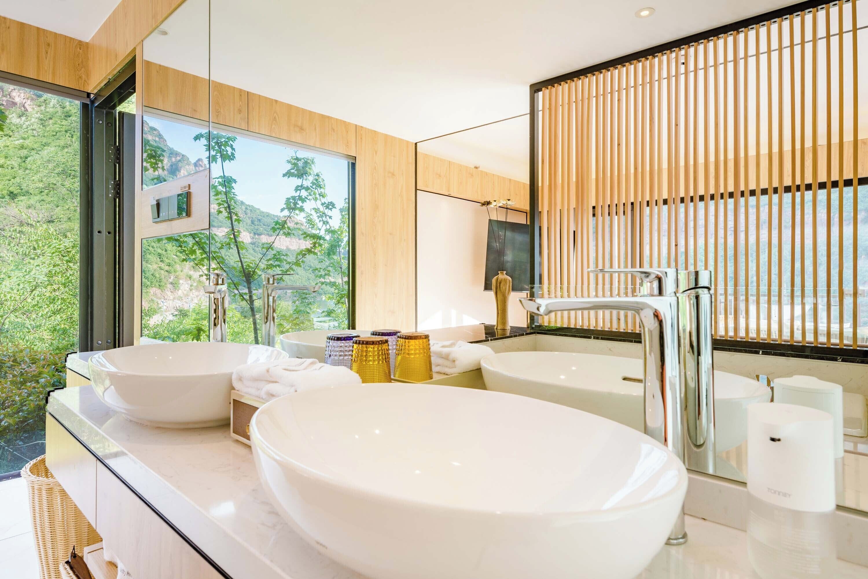 A modern bathroom with two large white sinks, a bathtub, and a large window offering a view of greenery and mountains.