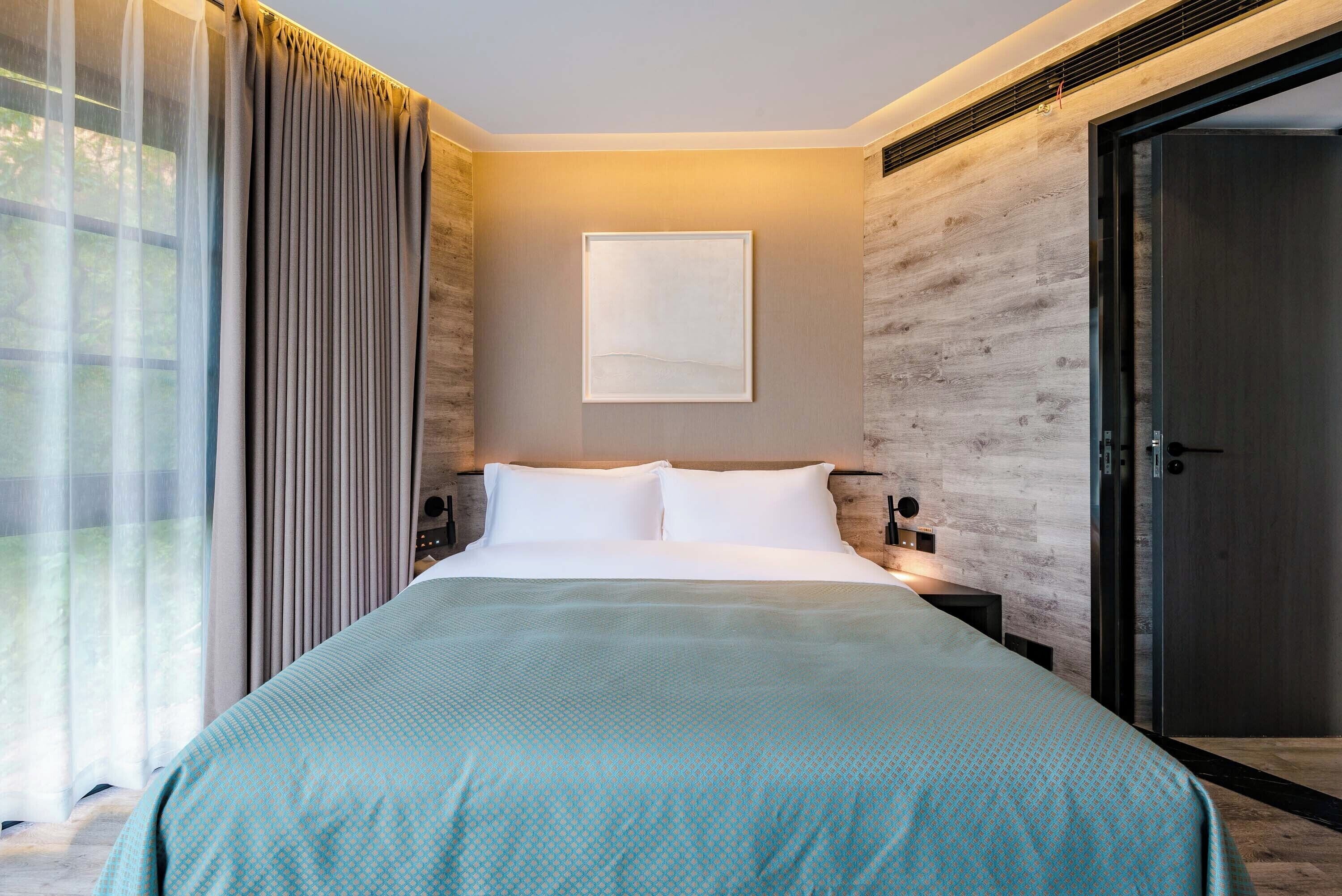 A modern hotel room with a large bed, a nightstand, a wall-mounted lamp, and a door.