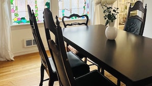Un comedor con una mesa y sillas de madera oscura, un florero blanco con flores, y dos ventanas con vidrieras de colores.