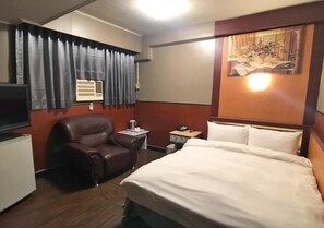 Ein Hotelzimmer mit einem Bett, einem Ledersessel, einem Schreibtisch mit Lampe, einem kleinen Tisch mit Telefon und einer Wand-Klimaanlage.