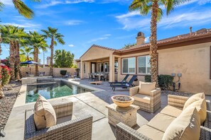 A pool area with lounge chairs and a dining table, surrounded by palm trees and a house with a tiled roof. - 4BR Indio Desert Escape • Stylish Stay • Mountain Views (Indio)