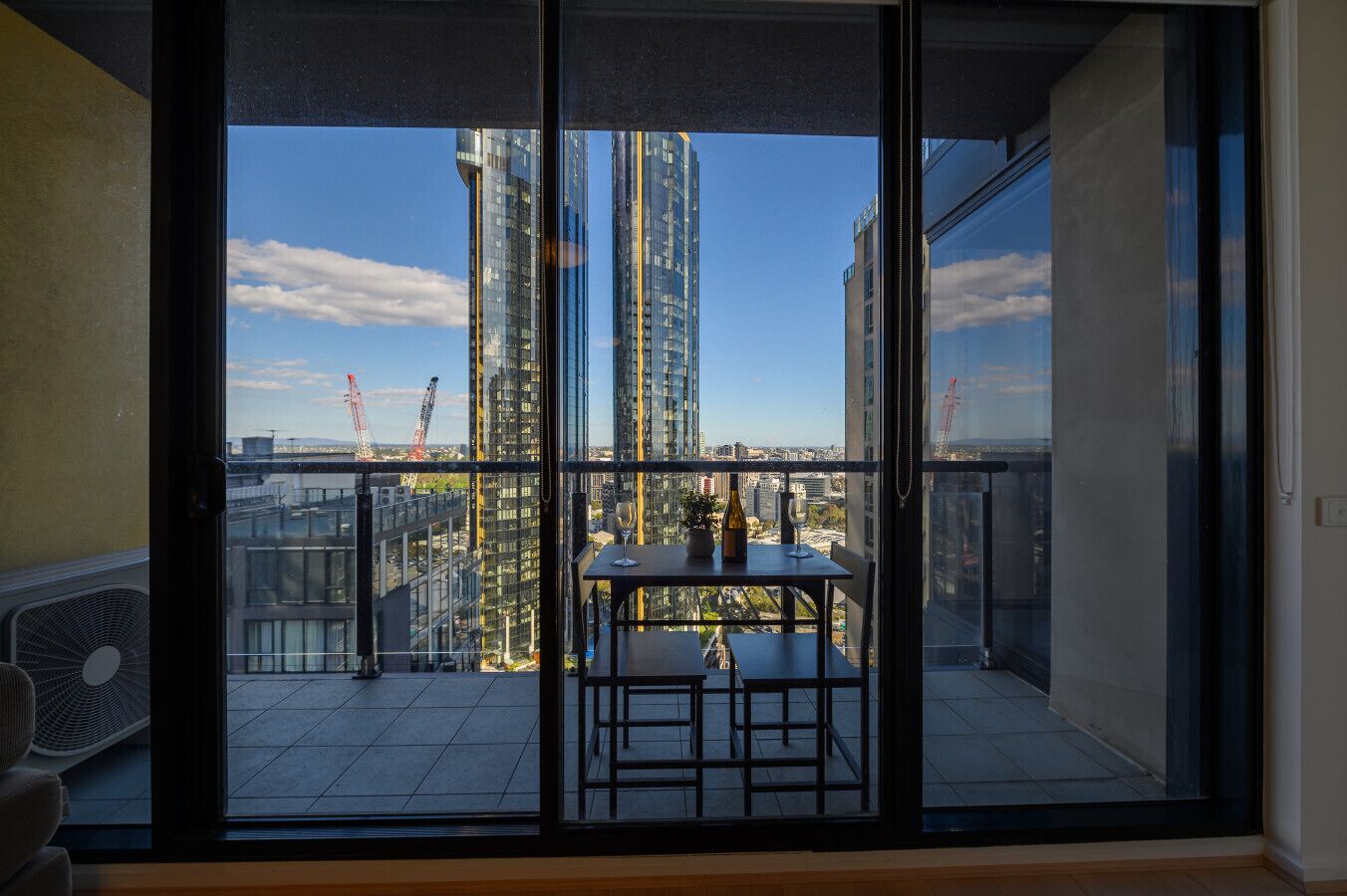 Un balcon avec une table et des chaises, un ventilateur, et une vue sur de hauts immeubles et des grues de chantier.