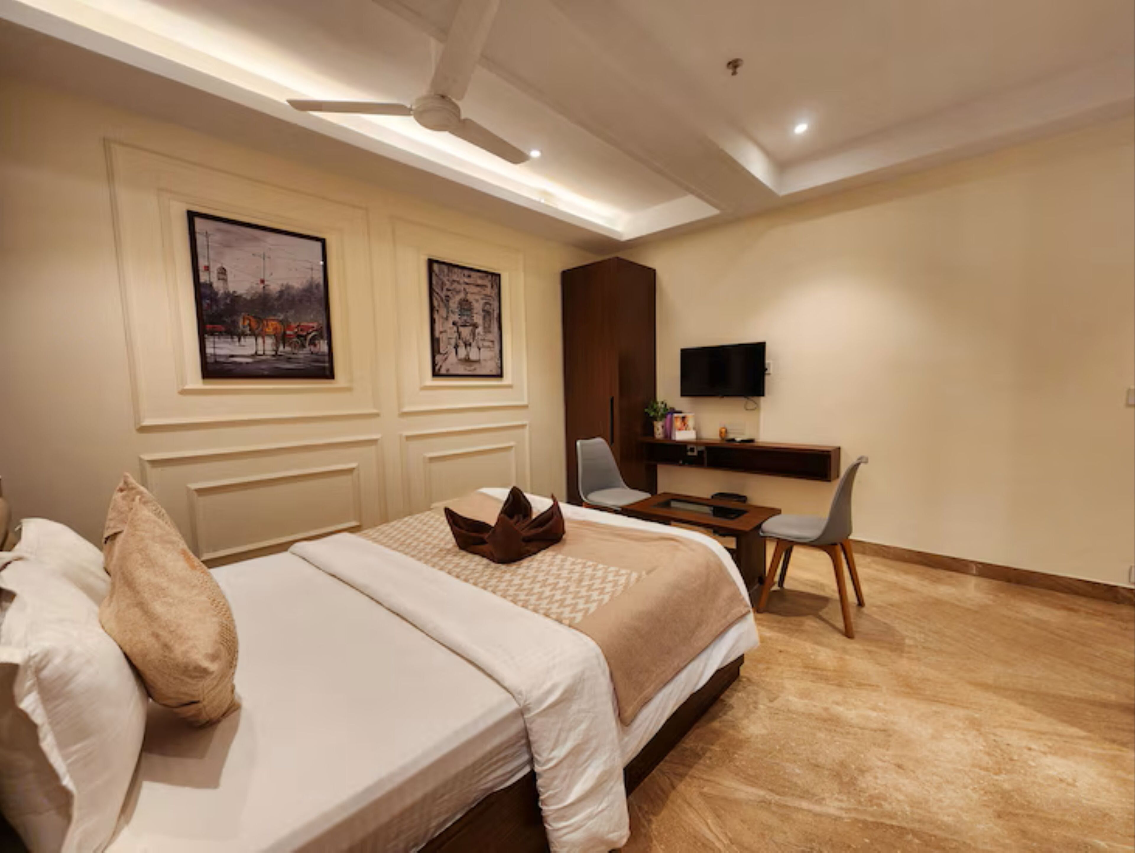A hotel room with a bed, a desk with a chair, a television, and two framed pictures on the wall.