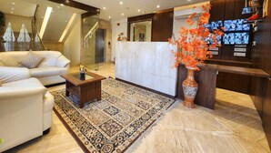A modern hotel lobby with a white sofa, a patterned rug, a reception desk, and a decorative vase with orange flowers. - EENNRA ARAMMBAGH (Arambag)