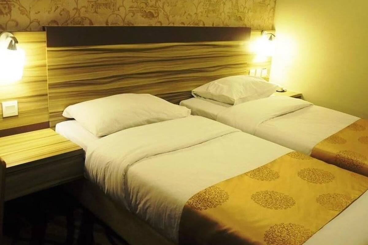 Habitación de hotel con dos camas, cada una con ropa blanca y un plaid decorativo dorado. Hay lámparas de noche y una mesita de noche con lámpara.