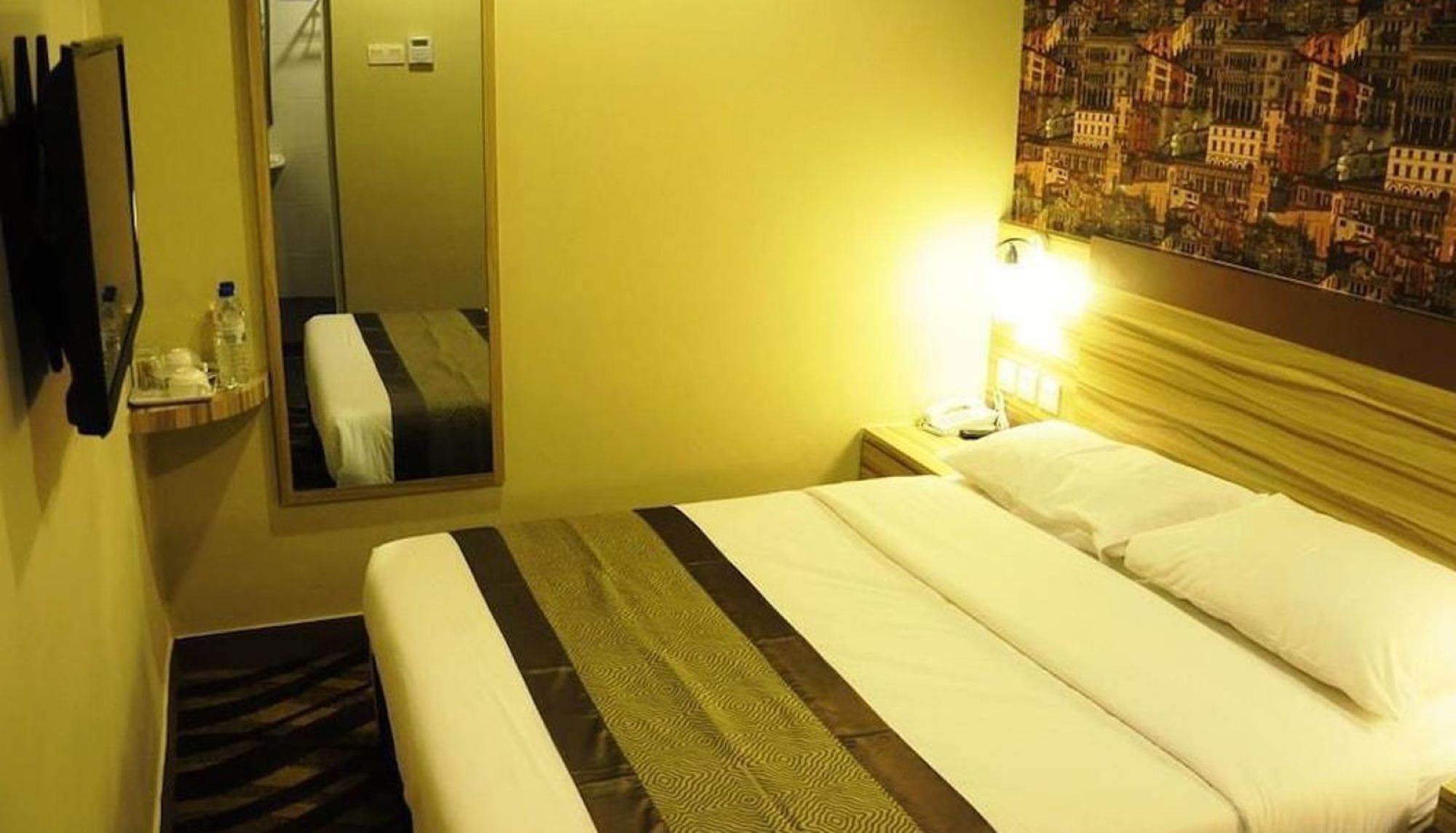 A hotel room with a bed, a bedside table with a lamp, a mirror, and a wall with a cityscape mural.