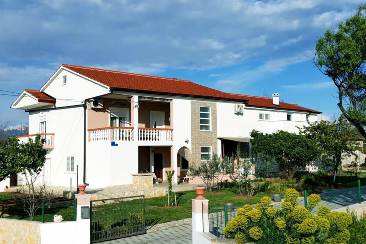 A two-story house with a red-tiled roof, a balcony, and a garden.