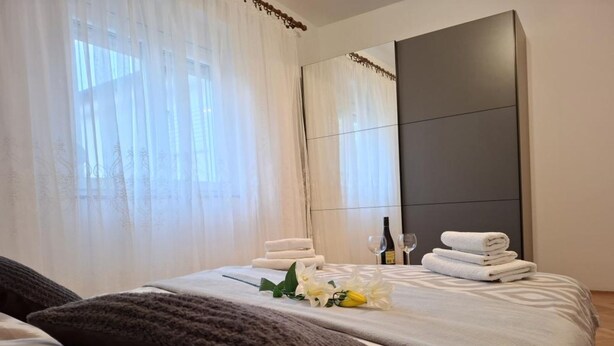 A hotel room with a large bed, a dark headboard, and a window with sheer curtains. There are towels and a bottle of wine on the bed. - Apartments Tomislav (Vrsi)