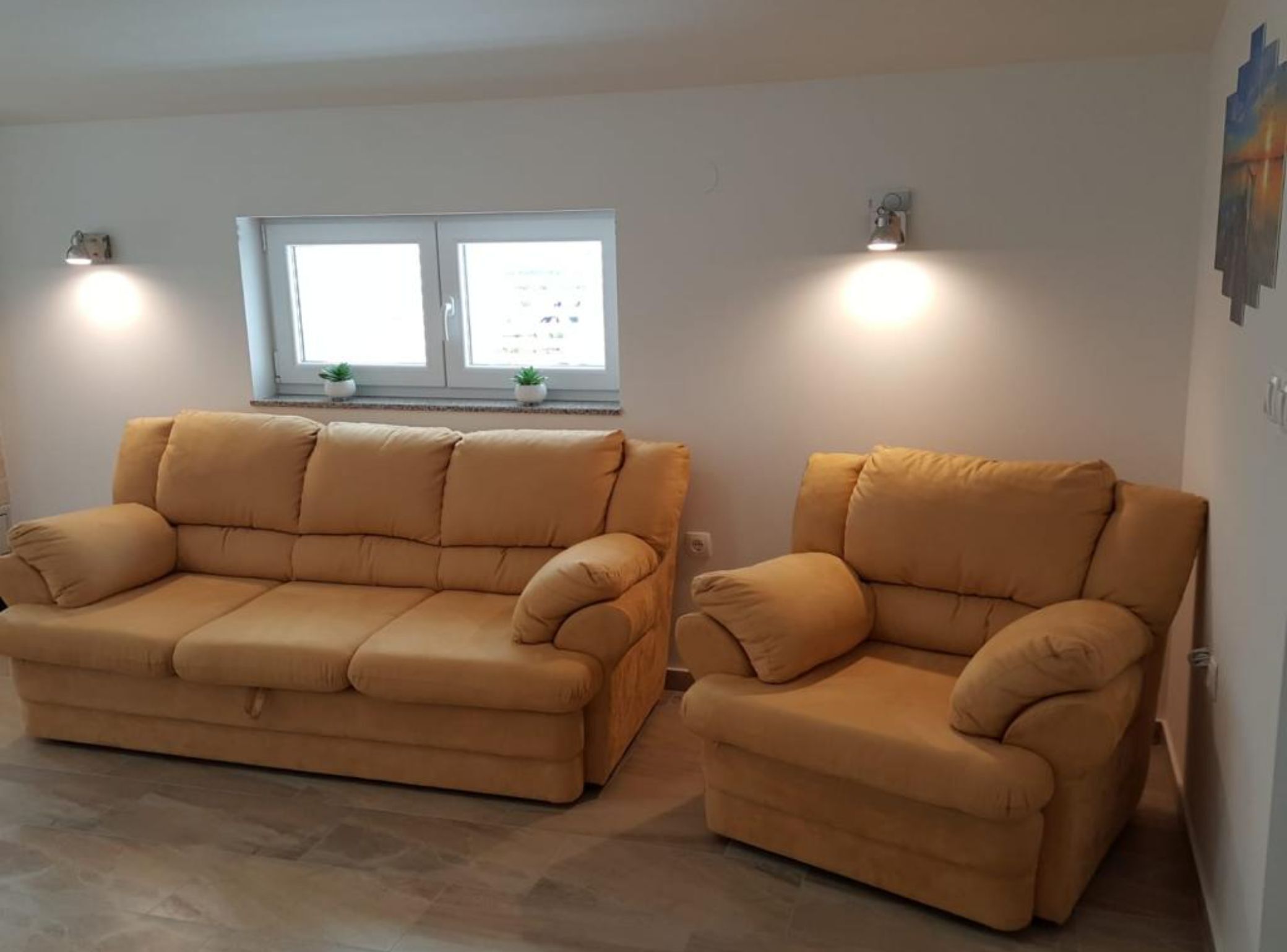 A living room with a beige sofa and armchair, a window with blinds, and two wall-mounted lights.