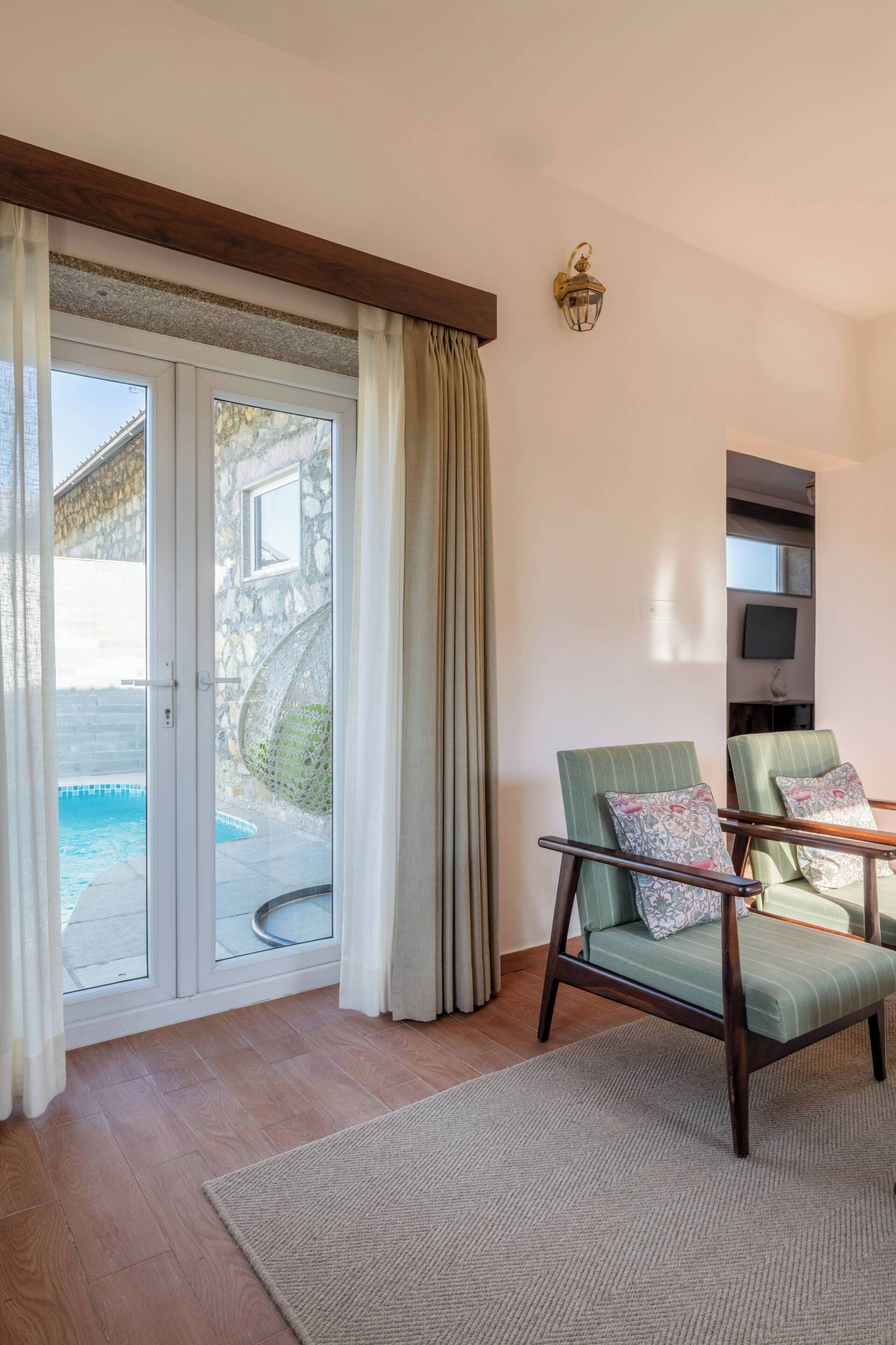 A room with a sliding glass door leading to a pool, a green upholstered chair with a patterned pillow, a wooden floor, and a wall-mounted TV.