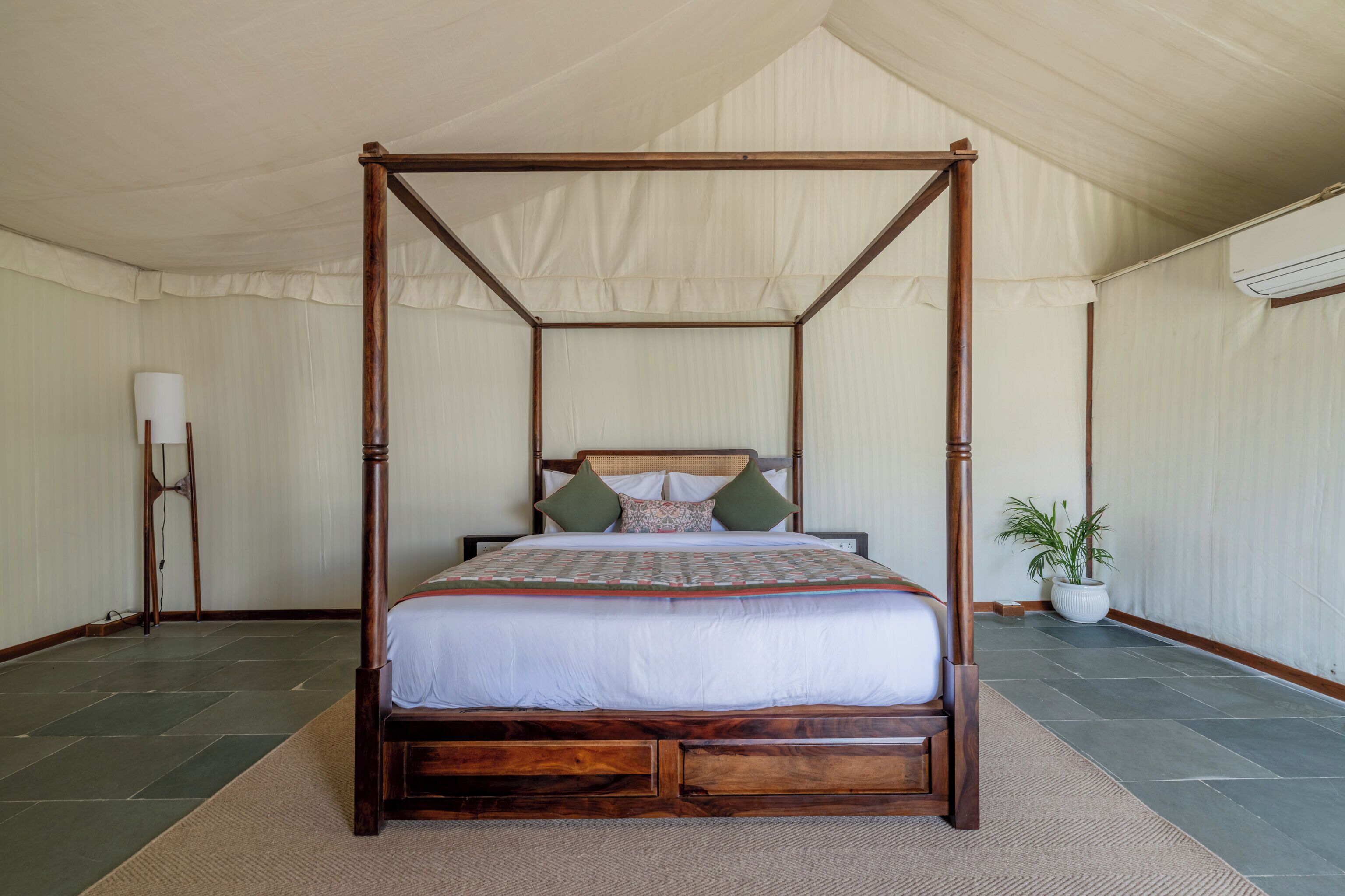 A four-poster bed with a wooden frame and white bedding, a potted plant, and a standing lamp in a tented room.