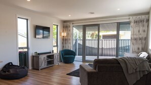 Un salon moderne avec un canapé, un fauteuil bleu, un téléviseur et une porte-fenêtre coulissante donnant sur un balcon.