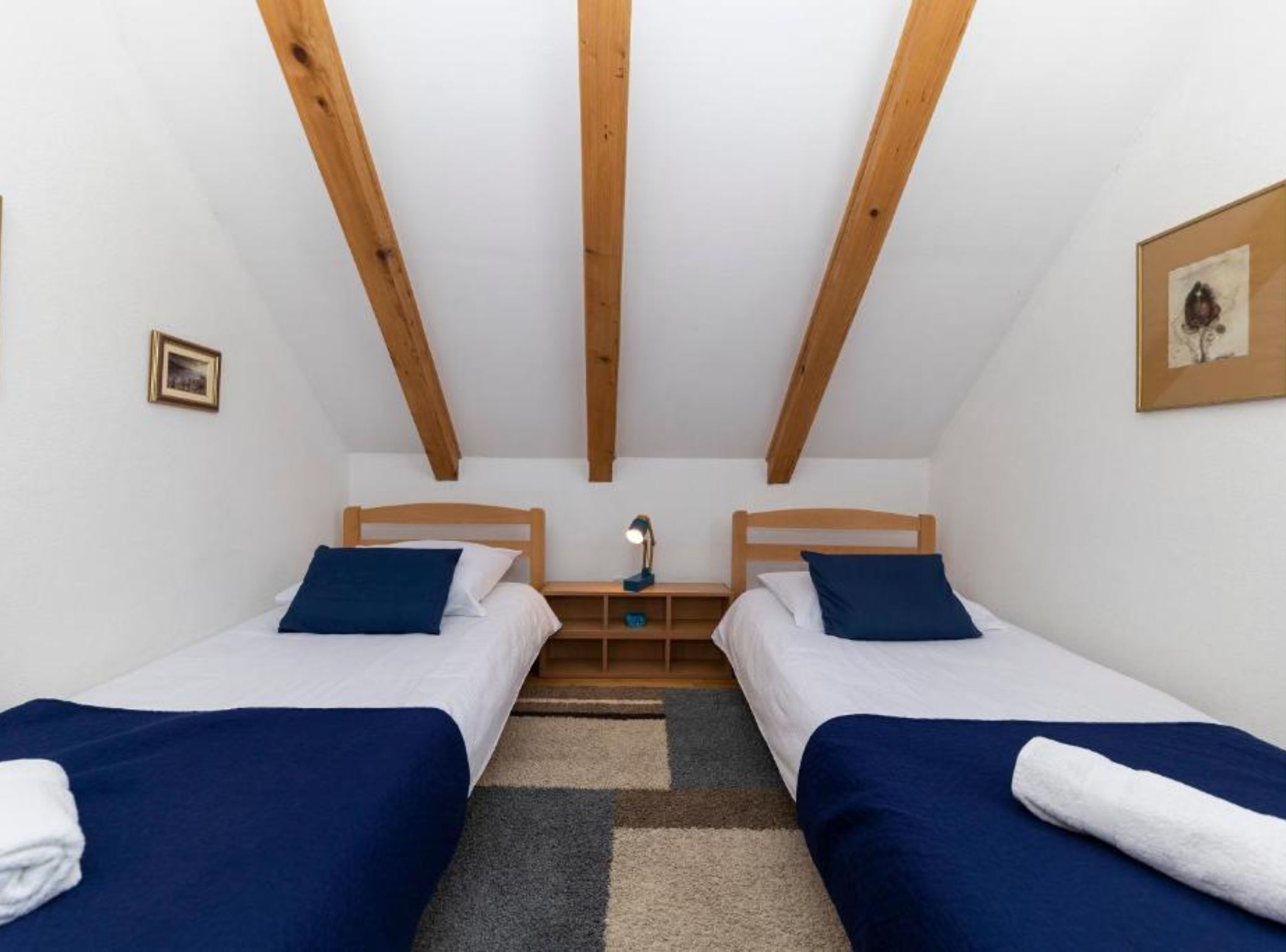 A small, cozy room with two single beds, wooden beams on the ceiling, a nightstand with a lamp, and a framed picture on the wall.