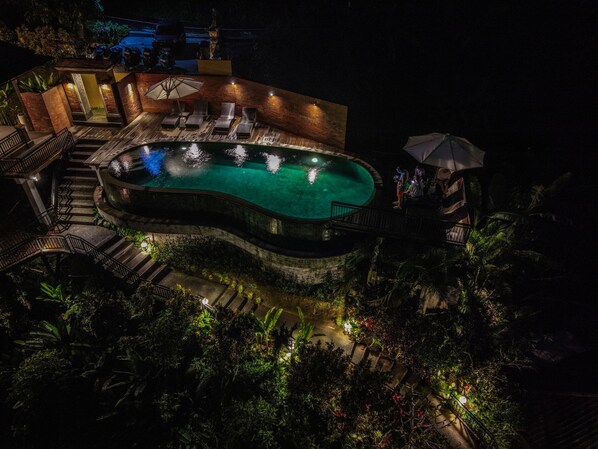 A nighttime view of a swimming pool with a wooden deck, lounge chairs, and a small pavilion. - The Tirta Sari Villas (Tegallalang)