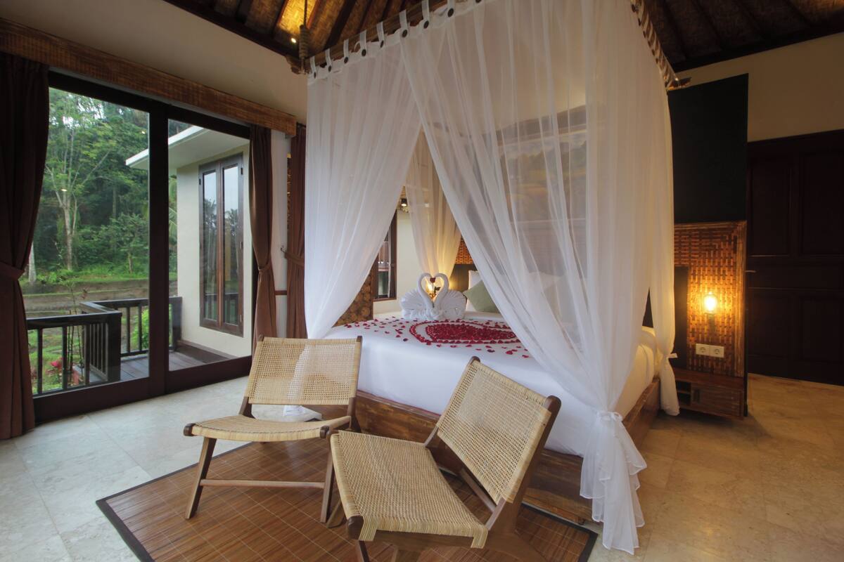 Une chambre à coucher avec un lit à baldaquin, deux fauteuils en osier, une grande fenêtre et une table de chevet avec une lampe.