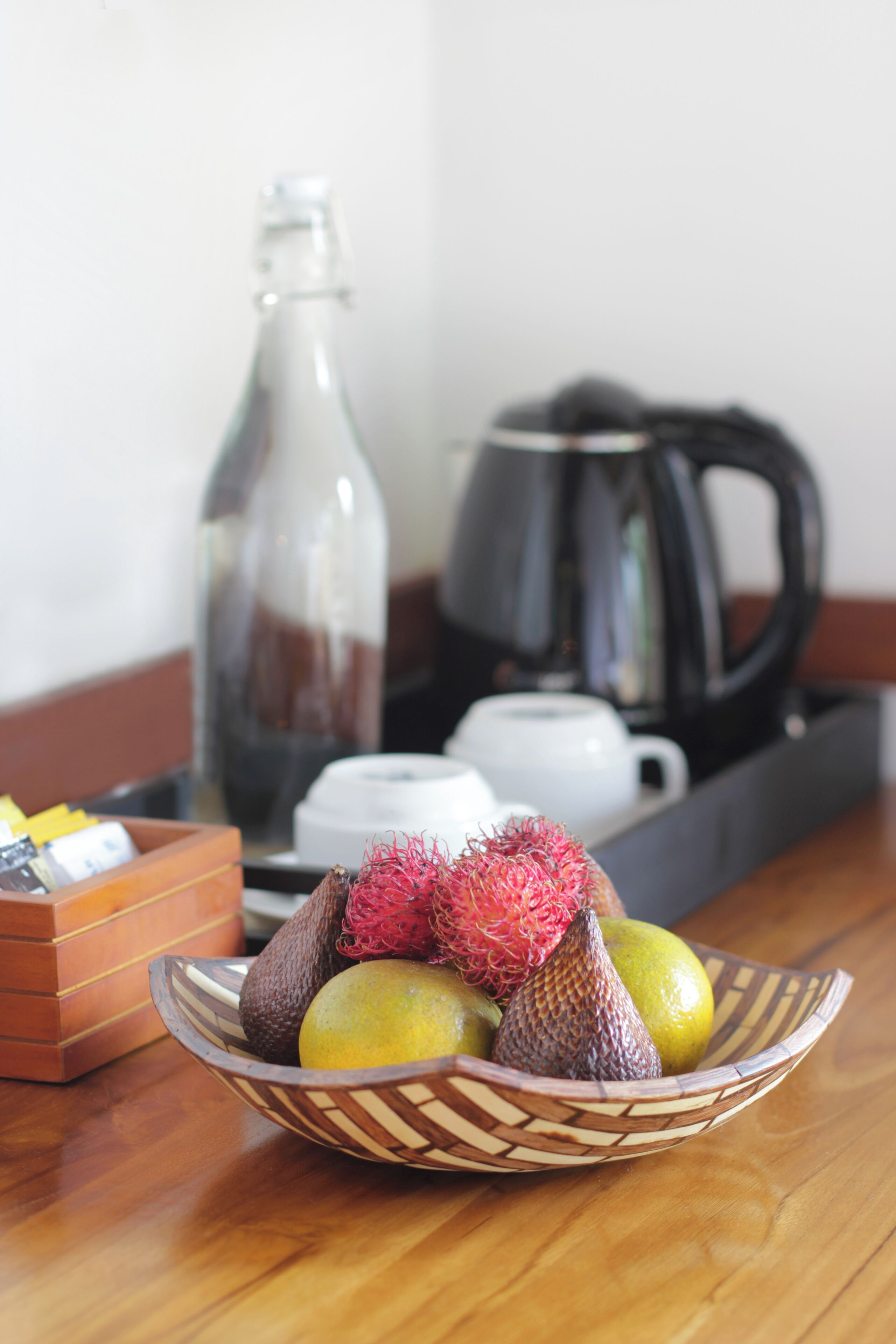 Une table en bois sur laquelle repose un panier en osier rempli de fruits, notamment des pommes, un fruit du dragon et un citron. On aperçoit, en arrière-plan, une machine à café et une bouteille en verre.