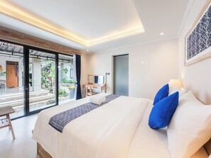 A modern hotel room with a large bed, a desk, and a view of the garden through the sliding doors. - Nandana Guest House Ubud (Ubud)
