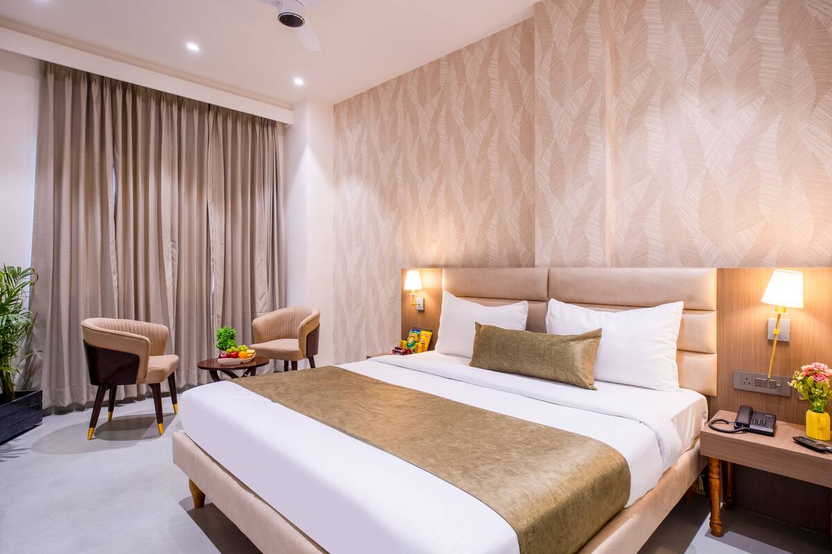A hotel room with a large bed, a bedside table with a lamp, a chair, and a small table with fruits.