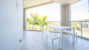 En balkon med hvidt bord og bænk, store vinduer og udsigt til grønt og bygninger.