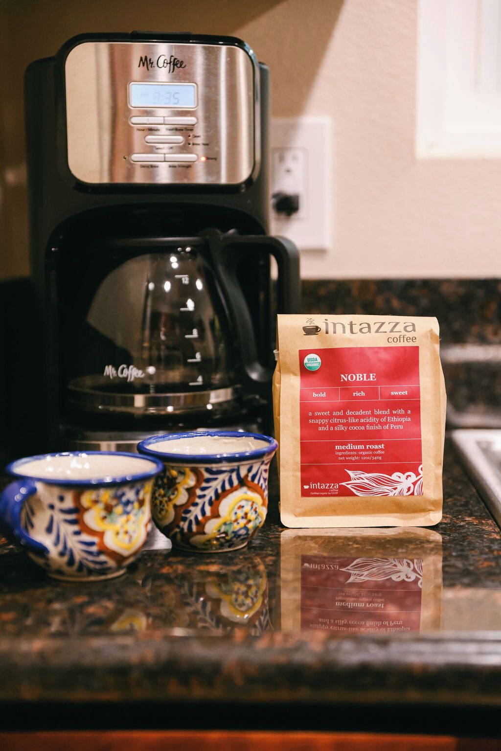A Mr. Coffee coffee maker, two ceramic cups, and a bag of Intazza coffee on a kitchen counter.