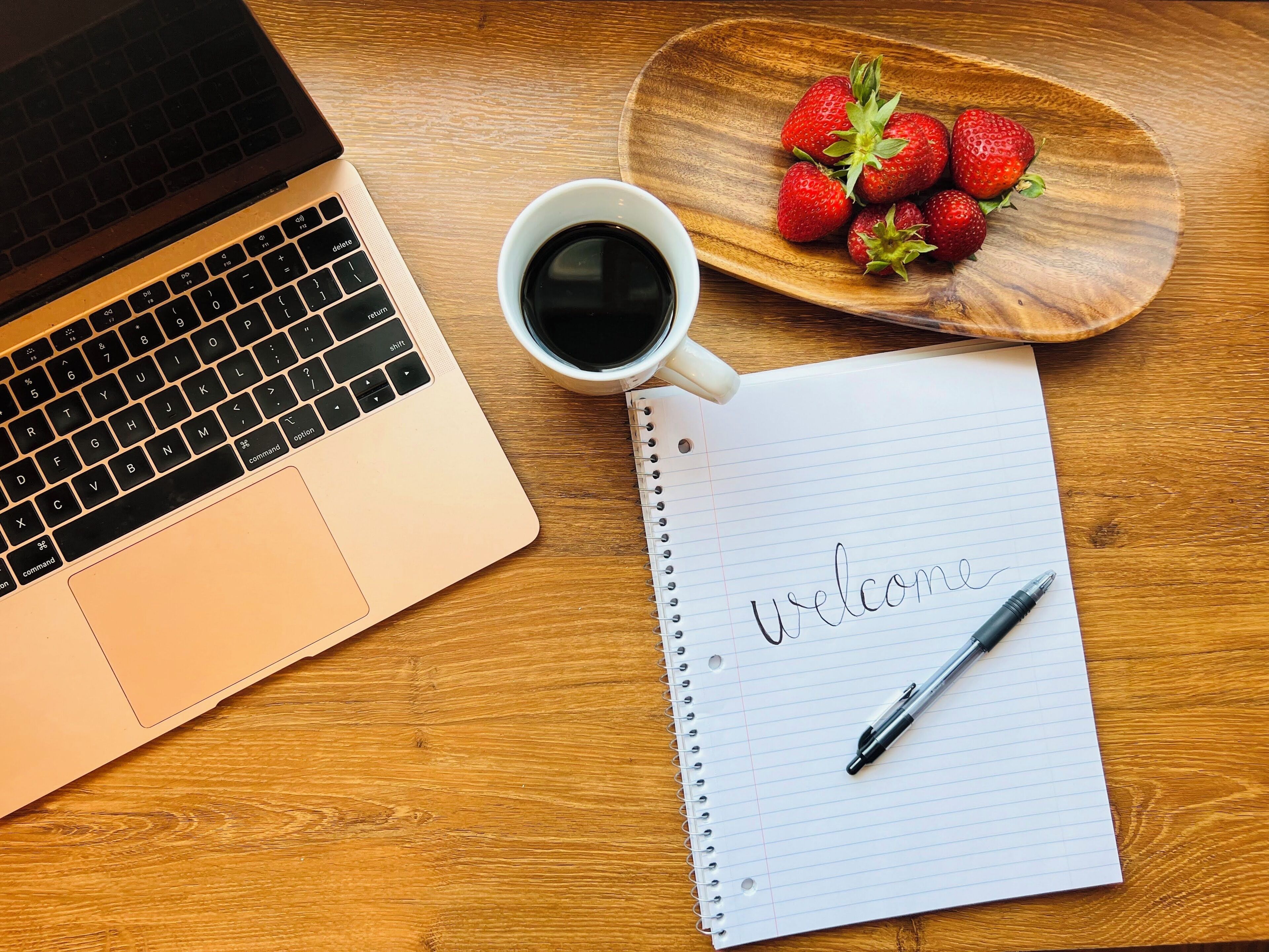 Ein Laptop, eine Tasse Kaffee, eine Schale Erdbeeren und ein Notizbuch mit dem Schriftzug 'Welcome'.