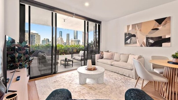 A modern living room with a large sliding glass door leading to a balcony, a round coffee table, a sofa, and a dining area with chairs. - Skyline Views & Pool | 2 King Beds, 2 Baths | Walk to South Bank | Free Parking (South Brisbane)