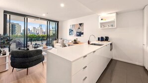 A modern kitchen with a large island, white cabinetry, and a view of the city through large windows. - Skyline Views & Pool | 2 King Beds, 2 Baths | Walk to South Bank | Free Parking (South Brisbane)