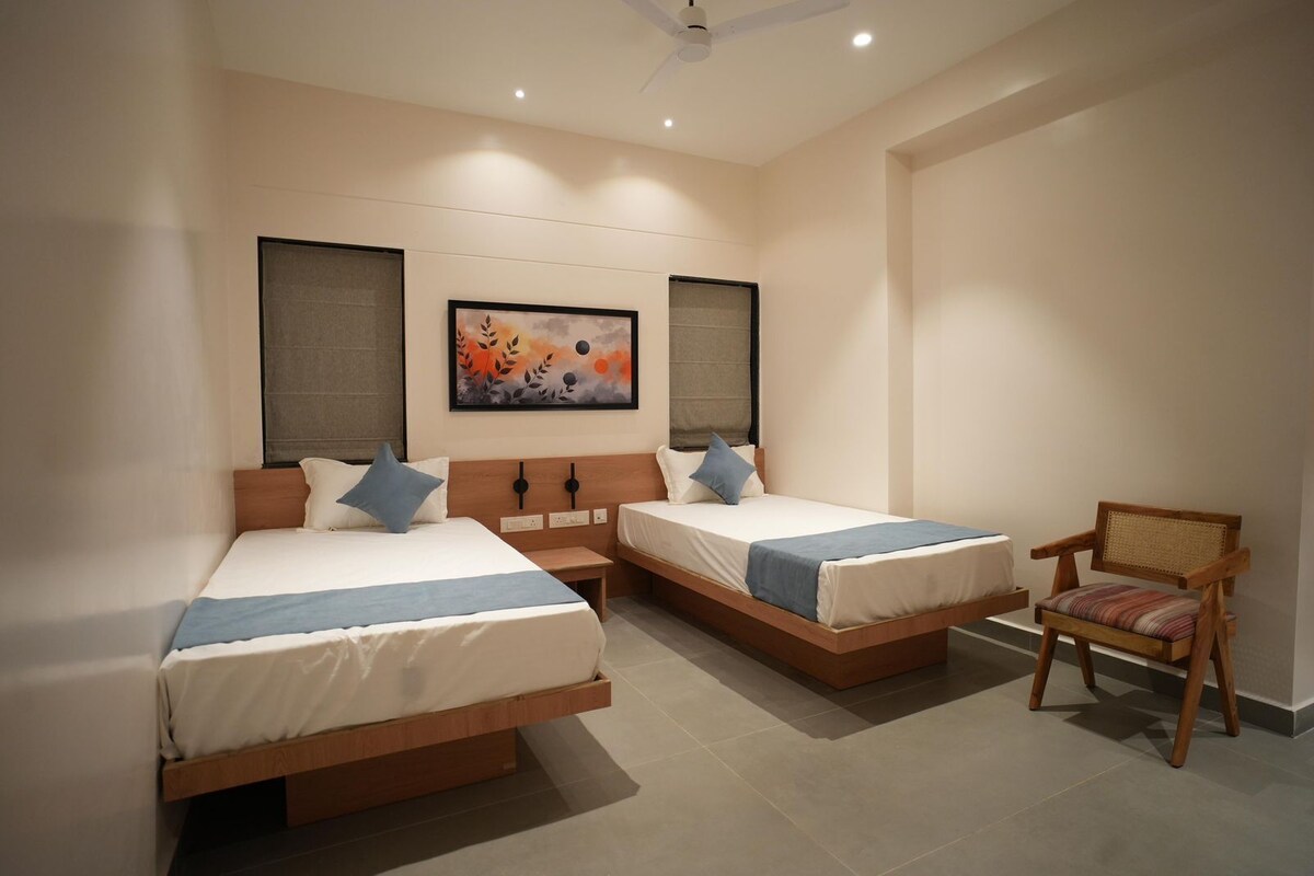 Una habitación de hotel con dos camas individuales, una silla de madera y obras de arte en la pared.