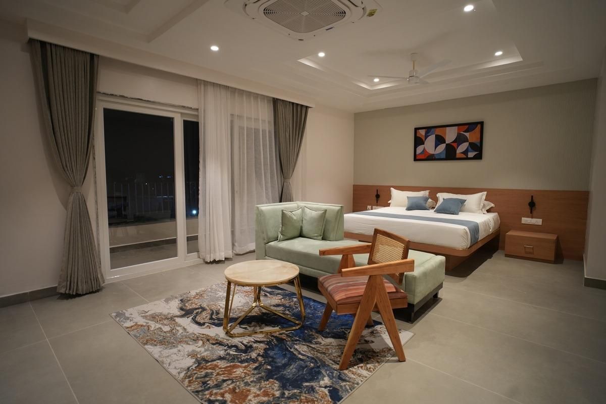 Habitación de hotel moderna con cama, sofá, sillón, una mesita y vistas a la ciudad de noche.