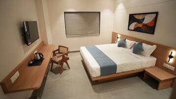 A modern hotel room with a wooden bed, a desk with a kettle, a chair, a small table, and a TV mounted on the wall.