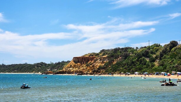 A beach with clear waters, a rocky cliff, and a forested hillside.