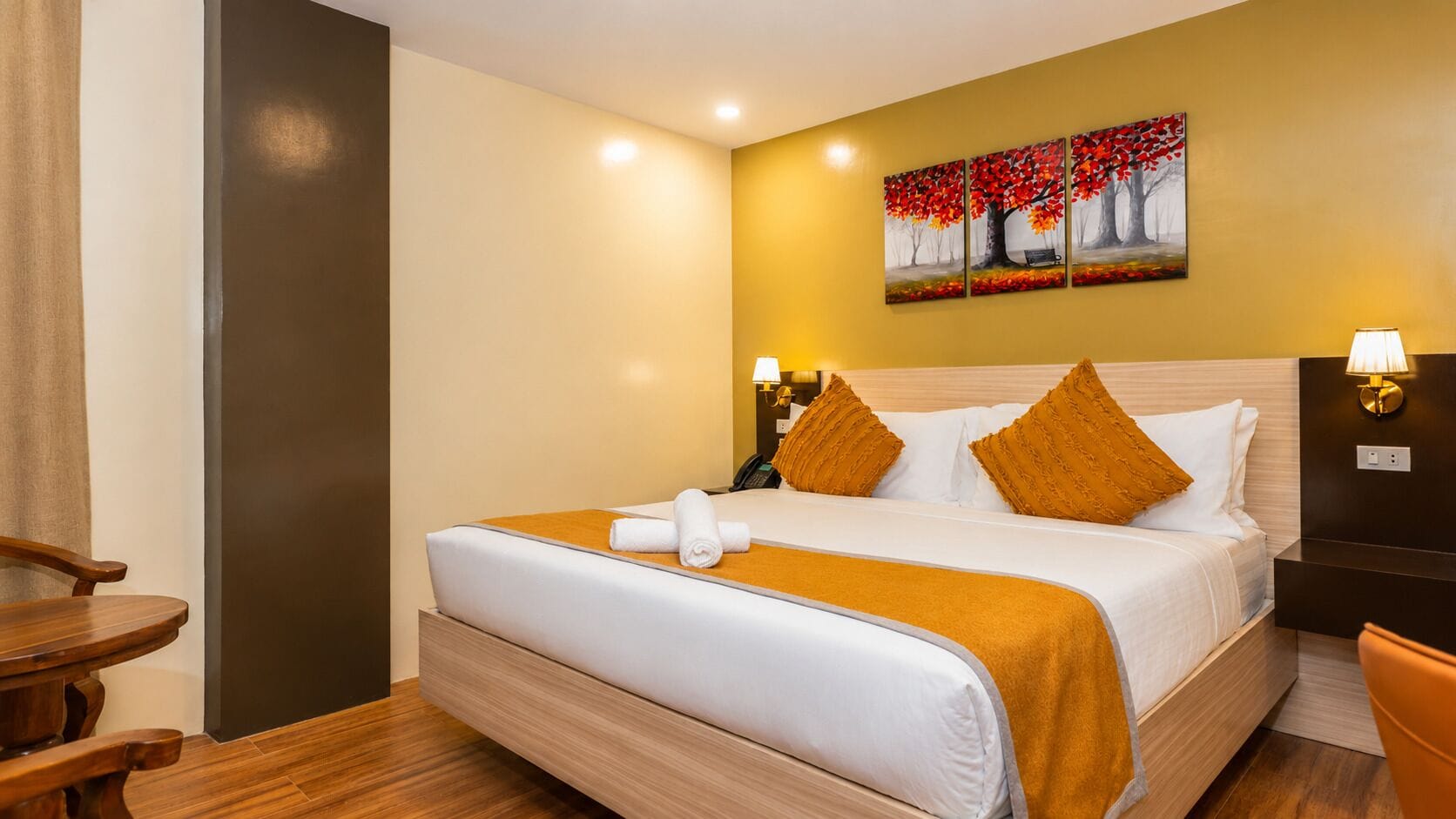 A hotel room with a bed, bedside lamps, a painting on the wall, and a wooden chair.