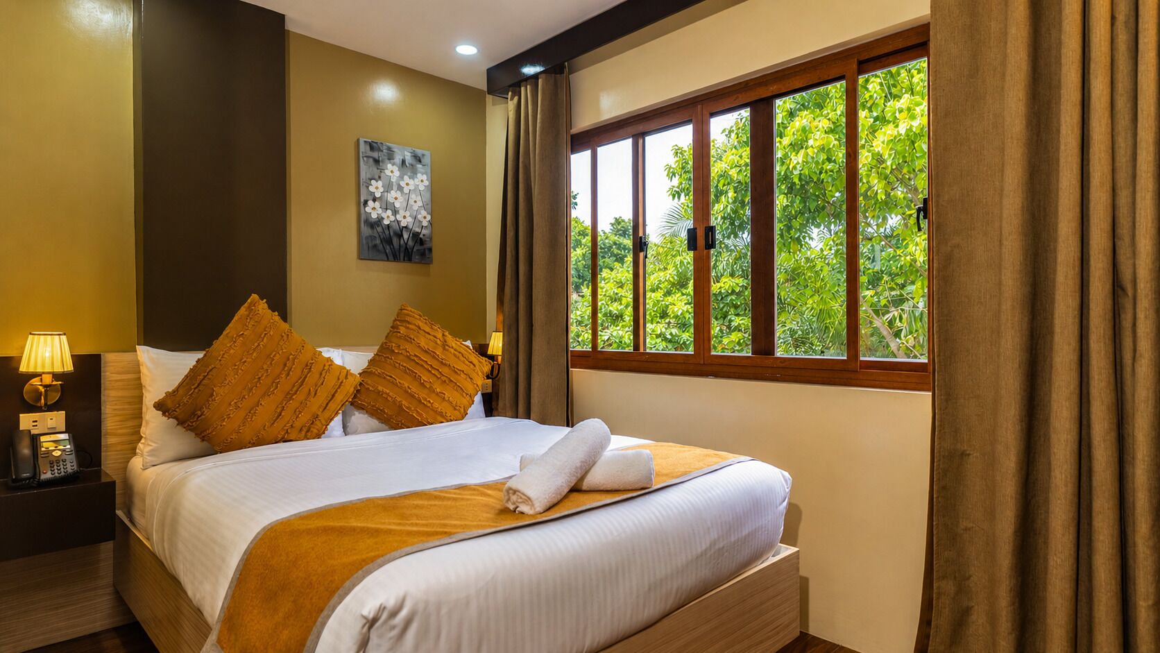 A hotel room with a large bed, a nightstand with a lamp, a painting on the wall, and a window with curtains.
