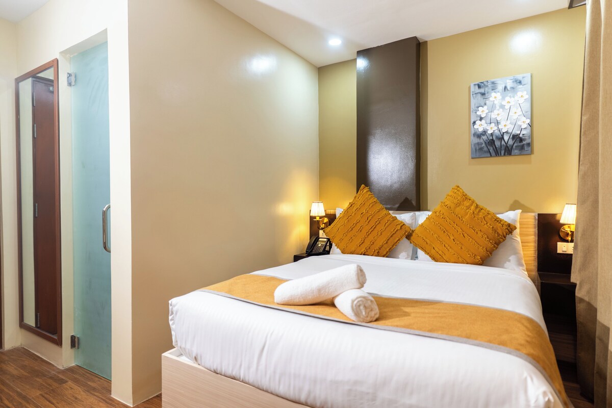 Une chambre d’hôtel comprenant un lit, deux tables de chevet, une petite table, un téléphone fixé au mur et une porte.