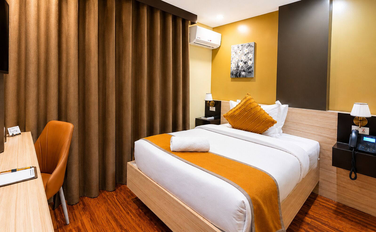 A hotel room with a bed, a desk, a chair, a nightstand, and an air conditioning unit.