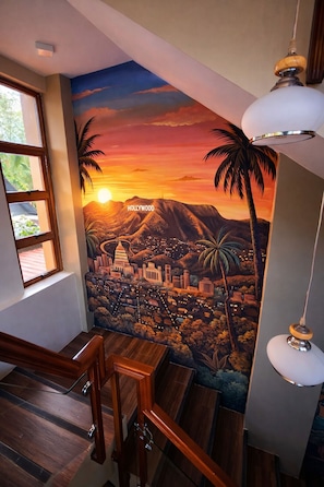 Un mural de un paisaje urbano al atardecer, con palmeras y la palabra 'Hollywood' en una escalera.