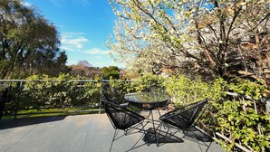 Un patio al aire libre con una mesa redonda y dos sillas, rodeado de abundante follaje y árboles en flor.