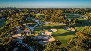 Vista aérea de un campo de golf con búnkeres de arena, árboles y un horizonte urbano distante.