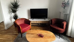Uma sala de estar com uma mesa de centro de madeira, duas poltronas vermelhas, uma televisão e um vaso de plantas.