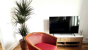 Uma sala de estar com uma poltrona vermelha estofada, uma grande planta em vaso, uma televisão de tela plana e um armário de madeira.