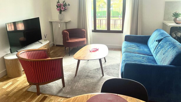 Uma sala de estar com televisão, sofás vermelhos e azuis, uma mesa de centro de madeira e uma cadeira vermelha.