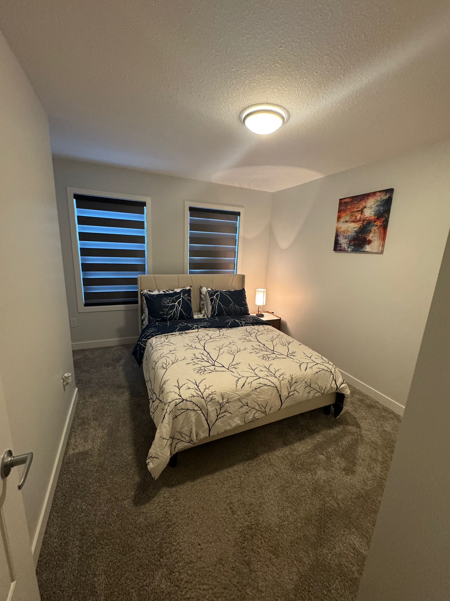 A bedroom with a bed, two windows with blinds, a ceiling light, and a painting on the wall.