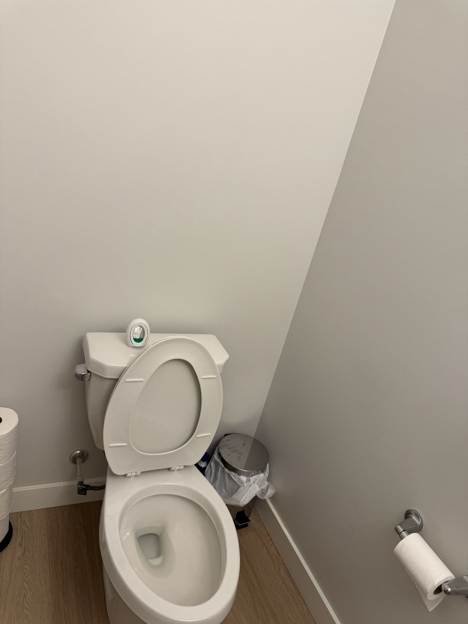 A white toilet with a closed lid, a toilet paper holder, and a trash can in a bathroom with a wooden floor.