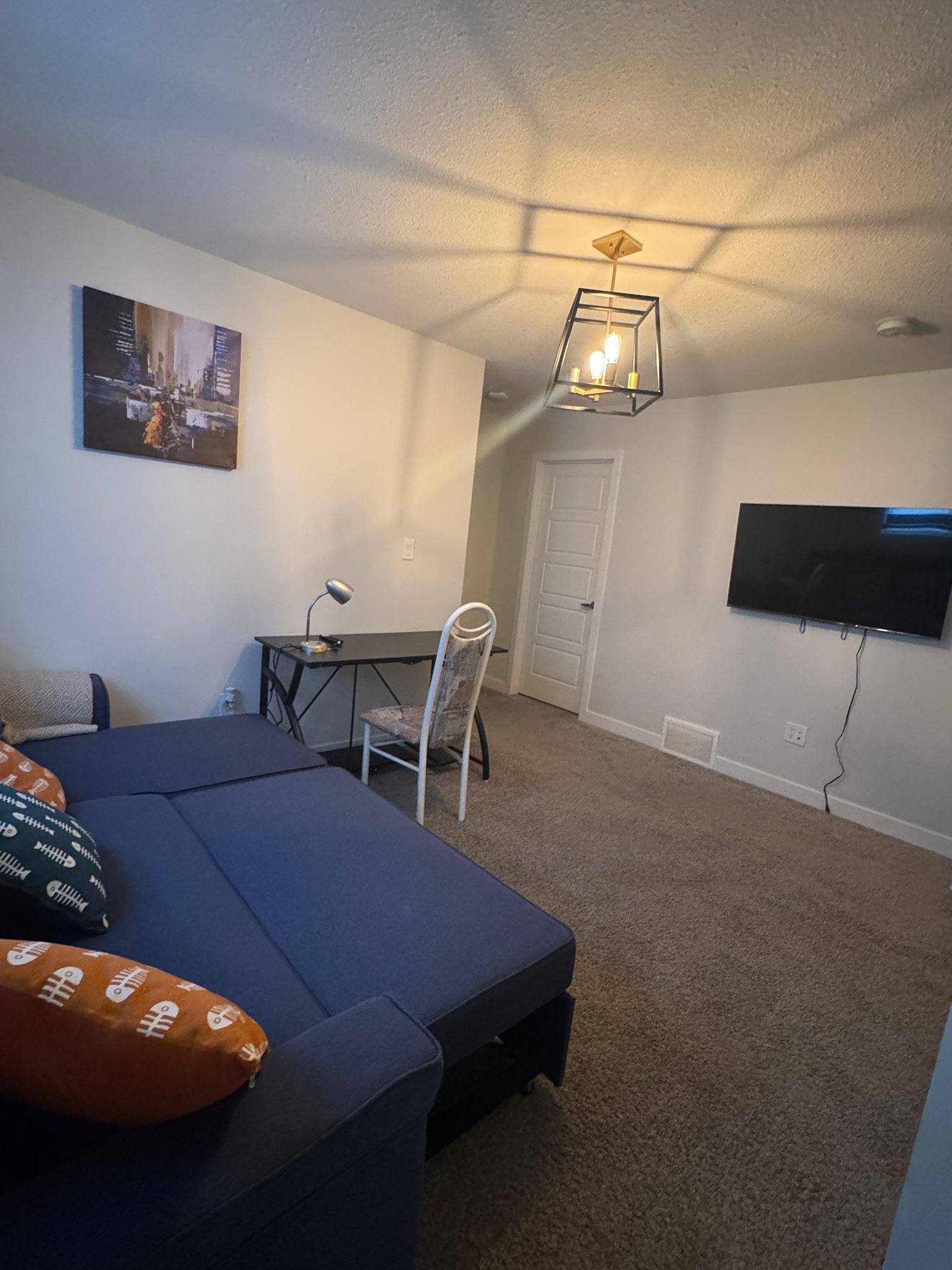 Une petite pièce bien éclairée, avec un lit, un bureau, une chaise, un téléviseur et un tableau accroché au mur.