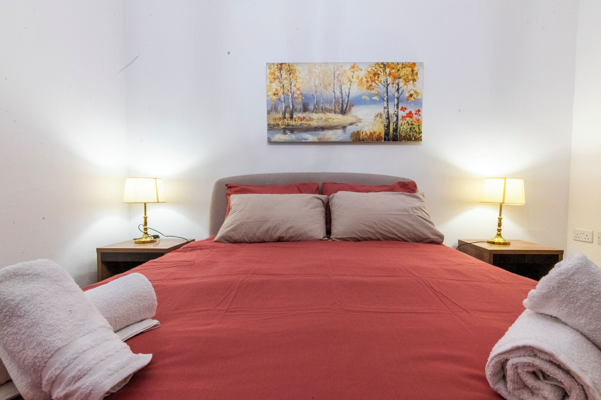 A neatly made bed with red bedding, two bedside lamps, and a painting of a landscape on the wall.
